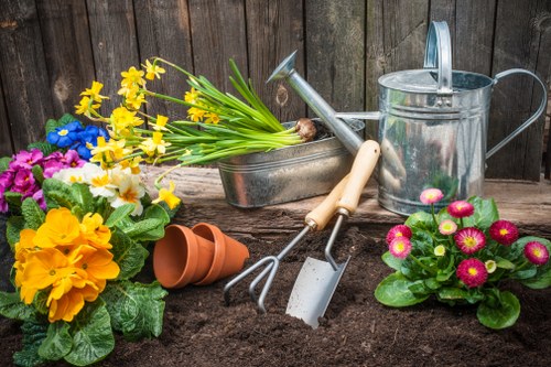 A vibrant garden scene featuring freshly turned dark soil in the foreground, surrounded by colorful flowering plants including yellow primroses, pink primulas, and white, purple, and yellow flowers, with various gardening tools such as a metal watering can, a hand rake, a trowel, and a small gardening fork placed on or partially inserted into the soil. Behind the tools, there are terracotta pots, some stacked, and a wooden fence serving as a backdrop. The overall setting appears to be a well-maintained outdoor garden space, with natural light illuminating the scene, suggesting a clear day suitable for gardening activities. This image highlights gardening and outdoor maintenance elements typical of services provided by Hedge Trimming Northolt in London, UK, supporting lush garden development and plant care.